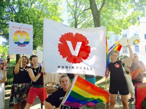 Prideparad 2015. I Rådhusparken, Hudik. Människor i V-tröjor och V-banderoll och prideflaggor.